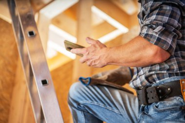 Bir inşaat işçisi, inşaat halindeki bir binanın parlak ışıklı bir bölgesinde iş eldiveni ve kot pantolon giyerken bir akıllı telefonu kontrol ederek merdivenin üzerinde duruyor..