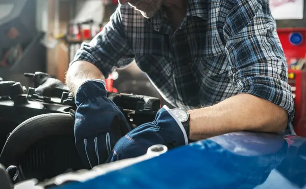 Bir insan garajda bir araba motorunu tamir etmeye odaklanır, mavi eldiven ve ekose gömlek giyer, otomotiv işinde kendini adamış ve yetenekli olduğunu gösterir..