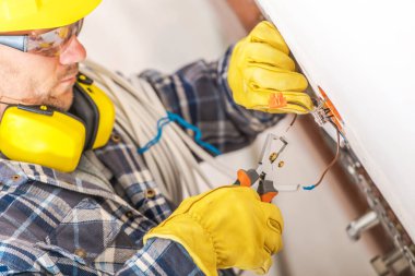 Yetenekli bir elektrikçi, koruyucu giysiler giyerken ve parlak bir konut ortamında özenle çalışırken elektrik kablolarını bağlamaya odaklanır..