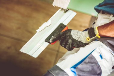 Bir insan ustalıkla duvar kenarına kürekle alçı sürüyor, kapalı alanda parlak bir şekilde aydınlatılmış eldiven giyiyor..