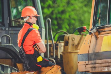 Kask takan ve yansıtıcı teçhizat kullanan bir inşaat işçisi, hareketli bir inşaat sahasındaki mola sırasında ağır bir makinenin üzerinde oturur..