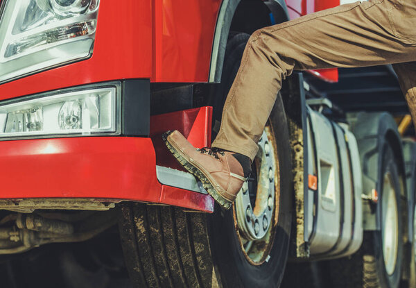 Человек в коричневых сапогах заходит в переднюю часть ярко-красного грузовика, припаркованного на транспортном складе в течение дня.