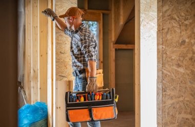 Bir inşaat işçisi ahşap bir duvarın yanında duruyor, elinde çeşitli aletlerle dolu bir alet çantası, inşaat halindeki bir evde çalışmaya hazırlanıyor..