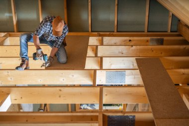 Bir marangoz, tahta kirişler ve güneş ışığıyla çevrili iken tahtaları korumak için elektrikli aletler kullanarak yeni bir eve döşeme döşemesini dikkatlice kurar..