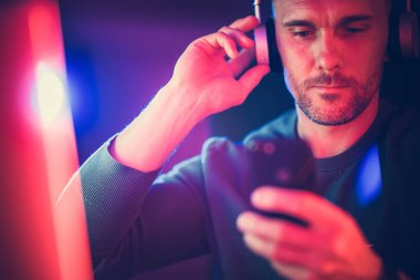 Kulaklıkla müzik dinlemeyi seven bir adam, renkli ışıklarla aydınlatılmış, karanlık bir ortamda akıllı telefonunu kontrol eder..