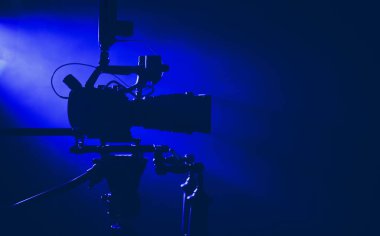 Bu kamera karanlık bir stüdyoda, canlı mavi ışıklarla aydınlatılmış bir tripodun üzerine yerleştirilmiş, bir film veya video yapımına hazırlanıyor..