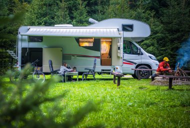 İnsanlar akşam ışığında kamp ateşinin sıcaklığının tadını çıkarırken, bir karavan yemyeşil bir alana park edilmiş..