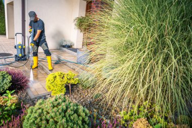 Sarı çizme ve şapka giyen bir adam, gün ışığında bahçedeki canlı bitkilerle çevrili seramik bir verandayı temizlemek için basınçlı çamaşır makinesi kullanıyor..