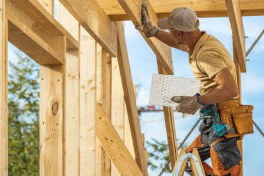 İnşaat işçisi, güneşli bir günde bir inşaat alanında merdivenin üzerinde dikilirken tahta kirişleri güvence altına almak için bir alet kullanıyor. Güvenlik ekipmanları görünür..