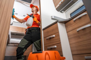 Turuncu gömlekli ve eldivenli bir işçi, bir karavanın içini sprey şişe ve bez kullanarak temizliyor..