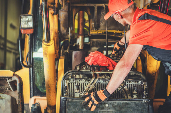 Механик в ярко-оранжевой одежде работает на двигателе желтой строительной техники внутри цеха, уделяя особое внимание ремонту и техническому обслуживанию.