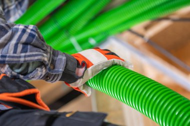 Bir işçi endüstriyel alandaki parlak yeşil boruları ayarlar, koruyucu eldivenler ve ekose gömlek giyer, malzeme taşırken güvenliği sağlar.