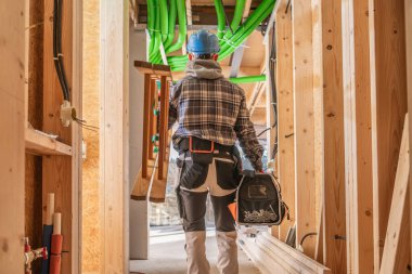 Bir inşaat işçisi, üzerinde yeşil borular bulunan ahşap bir iskelette yürürken, etkin bir inşaat alanının altını çizerek araçlar taşır..