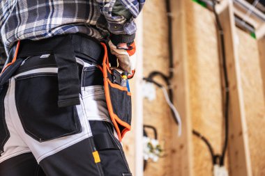 Bir inşaat işçisi, bir atölyede alet kemeri ve ekose gömlek giyerek ayakta duruyor ve elindeki aletlerle inşaat faaliyetlerine girişmeye hazır..