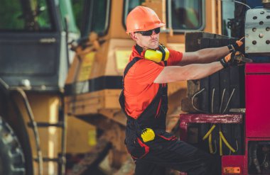 Turuncu kıyafetli bir inşaat işçisi, gün boyunca makine ve malzemelerle dolu yoğun bir şantiyedeki bir kamyona araç yüklüyor..
