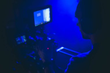 Bir kamera operatörü ekipmanını loş mavi ışıkta ayarlıyor, gece yaratıcı bir üretim ortamında çekim yapmaya odaklanıyor..