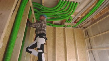 Bir işçi, elektrik ya da su tesisatı için havalandırma borusu döşemek için inşaat duvarına tırmanıyor. Dekor kısmen inşa edilmiş, ahşap çerçeveler sergileniyor..