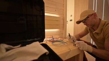 Şapkalı bir teknisyen şık, minimalist bir odadaki elektrik tesisatına dikkatle odaklanır. Cihazlar ve malzemeler yakınlarda organize edilir, dikkatli işçilik ve detaylara dikkat.