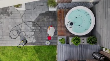 Bir profesyonel, dışarıdaki bir arka bahçedeki jakuziyi çevreleyen teras alanını temizlemek için basınç makinesini kullanıyor. Süreç, açık ve güneşli bir günde gerçekleşir ve eksiksiz temizlik sağlanır..