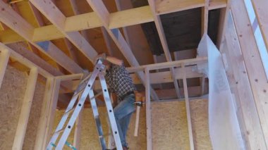 Bir inşaat işçisi yeni inşa edilmiş bir binaya tavan kirişleri yerleştirmek için merdivene tırmanır. Çalışma alanı ahşap yapılar ve tamamlanmamış duvarlarla dolu, aydınlık bir günde inşaat projesinin gidişatını gösteriyor..