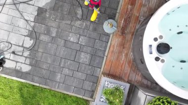 Bir kişi teras alanını temizlemek için basınç makinası kullanıyor. Dekorasyonda ahşap döşeme, jakuzi ve düzgünce düzenlenmiş bitkiler yer alıyor. Bu etkinlik güneşli bir öğleden sonra, açık hava bakım çalışmalarını vurguluyor..