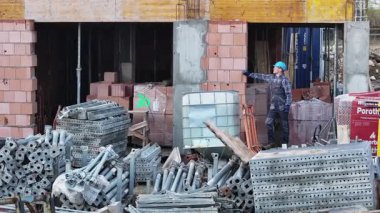 Bir inşaat işçisi işlek bir kentsel alanda materyalleri koordine ederken ve düzenlerken görülüyor. Bölge tuğla, boru ve metal yapı iskelesi yığınlarıyla dolu ve devam eden inşaat projesini sergiliyor..