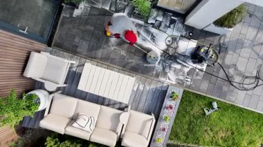 Bir kişi yeşil bitkiler ve açık hava mobilyalarıyla çevrili bir teras alanını temizlemek için basınç makinası kullanıyor. Su ve sabun spreyi fayansları kapladığında, şahıs koruyucu giysiler giyer ve parlak güneş ışığı altında temiz ve düzenli bir yüzeyi ortaya çıkarır..