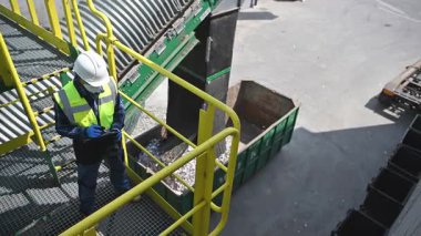 Güvenliğe saygılı bir işçi endüstriyel geri dönüşüm tesisinde bir platformda duruyor. Malzemeler taşıyıcıdan aşağıdaki büyük bir çöp konteynırına aktarılırken operasyonu izliyorlar. Çevre, temiz ve verimli bir geri dönüşüm sürecini yansıtıyor.