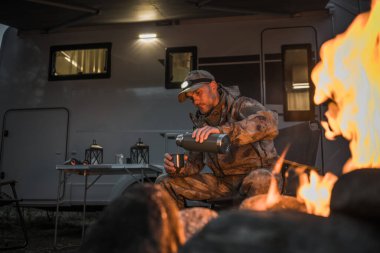 Açık hava kıyafetleri giyen bir adam kamp ateşinin yanında oturur ve bardağa sıcak bir içecek doldurur. Kamp kurulumunda arka planda bir minibüs ve yakınlardaki bir masada sıcak bir akşam atmosferi oluşturan fenerler var..