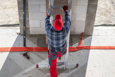 Kırmızı şapkalı ve eldivenli bir inşaat işçisi tuğla duvara ulaşmak için metal bir merdivene tırmanır. İşçi ekose gömlek ve kırmızı pantolon giyer, elimizdeki işe odaklanır..