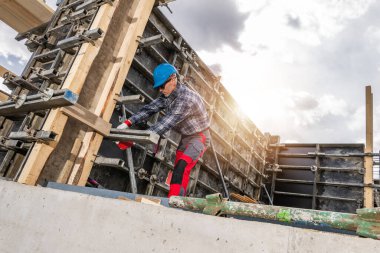 Mavi kasklı bir inşaat işçisi gün boyunca inşaat alanında beton döşeme hazırlıyor. Ağır ahşap kirişler ve çelik destekler, bulutların güneşi kısmen karartması nedeniyle dengeyi sağlamak için kullanılır..