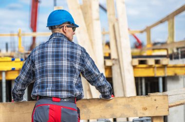Mavi bir şapka ve ekose gömlek giyen bir inşaat işçisi inşaat alanında devam eden çalışmaları izliyor. Arkaplanda parlak bir gökyüzünün altında tahta kirişler ve ağır makineler görülebilir..