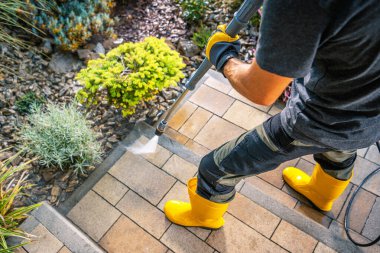 Bir insan, dışarıdaki basınçlı çamaşır makinesiyle taş merdivenleri temizliyor. Onlar merdivenlere su püskürtürken parlak sarı çizmeler ve siyah eldivenler giyiyorlar..