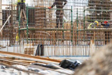 İnşaat işçileri, ekipman ve malzemelerle dolu bir şantiyede beton bir yapı inşa etmekle aktif olarak meşgul. Güçlendirme çubukları ve yapı iskelesi inşaatın ileri safhalarına işaret ediyor.