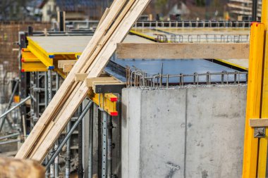 İnşaat alanında inşaat iskelesi tarafından desteklenen kısmen tamamlanmış beton duvarlar sergileniyor. Ahşap kirişler stratejik olarak yerleştirildi. İşçiler modern mühendislik tekniklerini vurgulayan bir sonraki aşamaya hazırlanıyorlar..