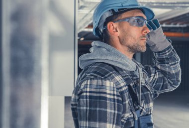 Miğfer ve güvenlik gözlüğü takan bir inşaat işçisi kısmen tamamlanmış bir binada duruyor. Yukarıya, derin düşüncelere bakıyor, etrafındaki yapıyı değerlendiriyor, işinin ciddiyetini vurguluyor..