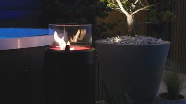 Açık Hava Akşam Toplantısı. Etrafı yumuşak ışıklandırmalı bitkilerle çevrili şık bir ateş kuyusu.