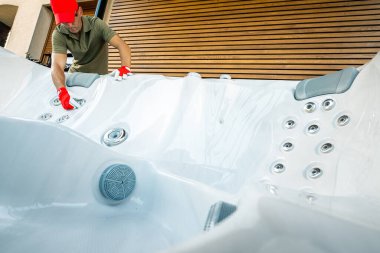 Kırmızı şapkalı ve eldivenli bir teknisyen, bir yerleşim bölgesinde jakuziyi titizlikle temizliyor. Parlak güneş ışığı küvetin parlak beyaz yüzeyini aydınlatır. Teknisyen, çeşitli jet ve filtreleri inceler ve korur..