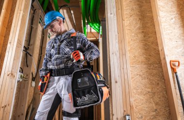 Bir inşaat işçisi, kısmen bitmiş bir binanın içinde, sert bir şapka ve güvenlik eldiveni giyerken görülüyor. Alet kemerini ayarlıyor ve vida ve gerekli ekipmanlarla dolu alet çantasına bakıyor..