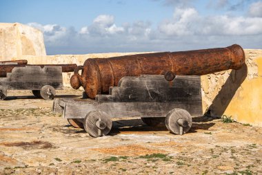 Paslı metalden yapılmış tarihi bir top bir kaledeki ahşap bir vagonda duruyor. Güneş ışığı sahneyi aydınlatır, altındaki desenli taş yüzeyi vurgular..