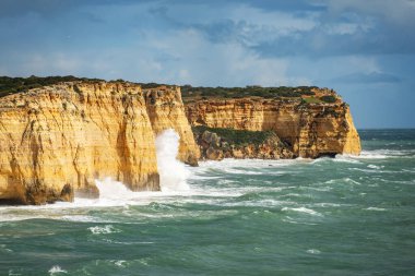 Kıyı şeridi boyunca, dalgalar dramatik sarı uçurumlara şiddetle çarparak beyaz köpük püskürtüyor. Okyanus koyu mavi-yeşildir, kaya çıkıntılarının sıcak tonlarıyla keskin bir tezat oluşturur..