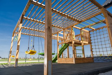 İyi tasarlanmış ahşap bir oyun parkında salıncaklar, bir kaydırak ve tırmanma duvarı çocukları oynamaya ve keşfetmeye davet ediyor. Yapı, üzerinde güzel açık mavi bir gökyüzü bulunan geniş bir eğlence alanına kurulmuştur..