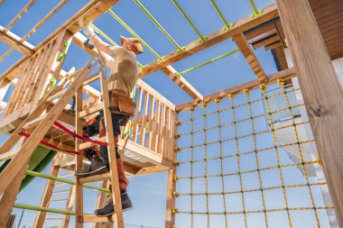 Bir inşaat işçisi, bir yerleşim bölgesinin arka bahçesindeki ahşap oyun yapısına dikkatlice güvenlik özellikleri yerleştirir. Parlak güneş ışığı, yeşil güvenlik halatlarını kullanarak merdivenleri çıkarken işçinin dikkatini çekiyor..
