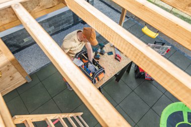 Bir inşaat işçisi, açık havada ahşap bir platformdaki alet çantasına çeşitli aletler yerleştirmeye odaklanır. İşçi kep takar ve çalışma alanına dağılmış diğer aletlerle çevrilidir. Bu yerleşim yeri güneş ışığıyla aydınlanır.,