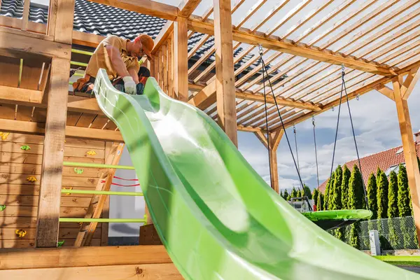 Bir adam dikkatle konutun arka bahçesindeki ahşap oyun yapısına parlak yeşil bir kaydırak yerleştiriyor. Oyun alanında tahta kirişler, tırmanma tutacakları ve salıncaklar çocuklara açık havada oynama olanakları sağlıyor. Ağaçlar yemyeşil.