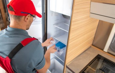 Bir teknisyen, çağdaş bir karavan mutfağında bakım sağlarken taze ürünleri buzdolabına koyarken görülüyor. Odaklanan işçi, ögeler için en uygun depolamayı sağlar.