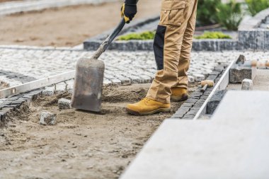 Sağlam giysiler içindeki bir inşaat işçisi bahçedeki patikaya taş kaldırımları dikkatlice yerleştiriyor. Proje açık gökyüzünün altındaki bir yerleşim bölgesinde gerçekleşiyor..