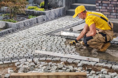 Sarı gömlekli ve kepli bir inşaat işçisi bahçeye bir yol açmak için kaldırım taşlarını titizlikle yerleştiriyor..