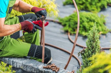 Bir peyzaj işçisi, yeşillik dolu canlı bir bahçede sulama borularını kesmek için budama makası kullanarak taştan bir çıkıntıda oturur. Güneş tepemizde ışıldıyor.