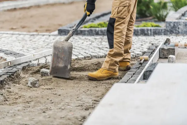 Sağlam giysiler içindeki bir inşaat işçisi bahçedeki patikaya taş kaldırımları dikkatlice yerleştiriyor. Proje açık gökyüzünün altındaki bir yerleşim bölgesinde gerçekleşiyor..